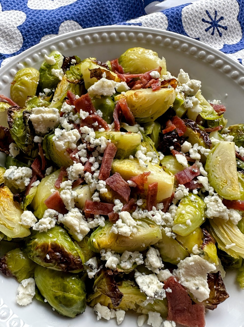 Roasted brussels sprouts in a white ceramic dish topped with bacon bits and blue cheese crumbles.