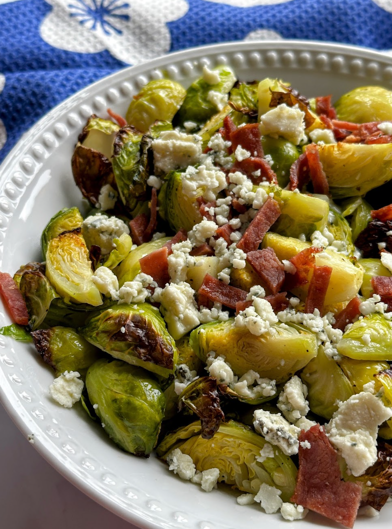 A rustic ceramic dish filled with golden roasted Brussels sprouts topped with crispy turkey bacon and blue cheese crumbles, lightly drizzled with dressing.
