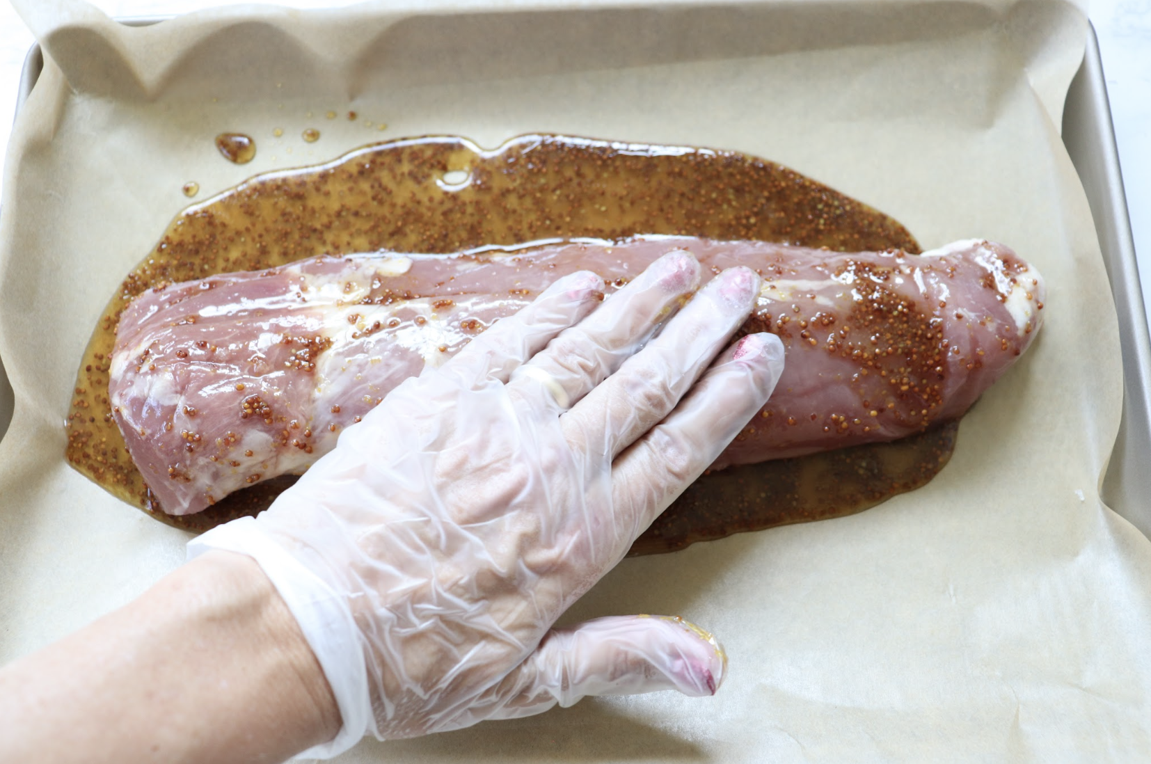 A pork tenderloin on parchment paper, topped with a maple glaze. A gloved hand is coating the pork.