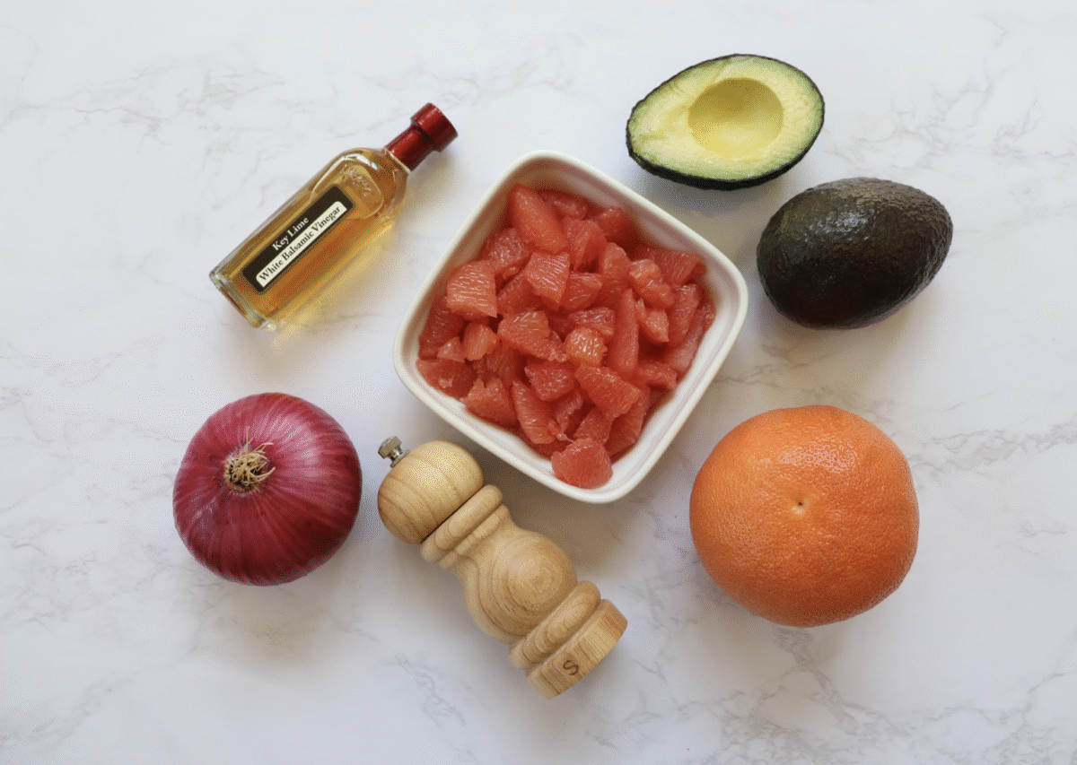 Ingredients needed to make citrus avocado salad set out on a marble countertop.