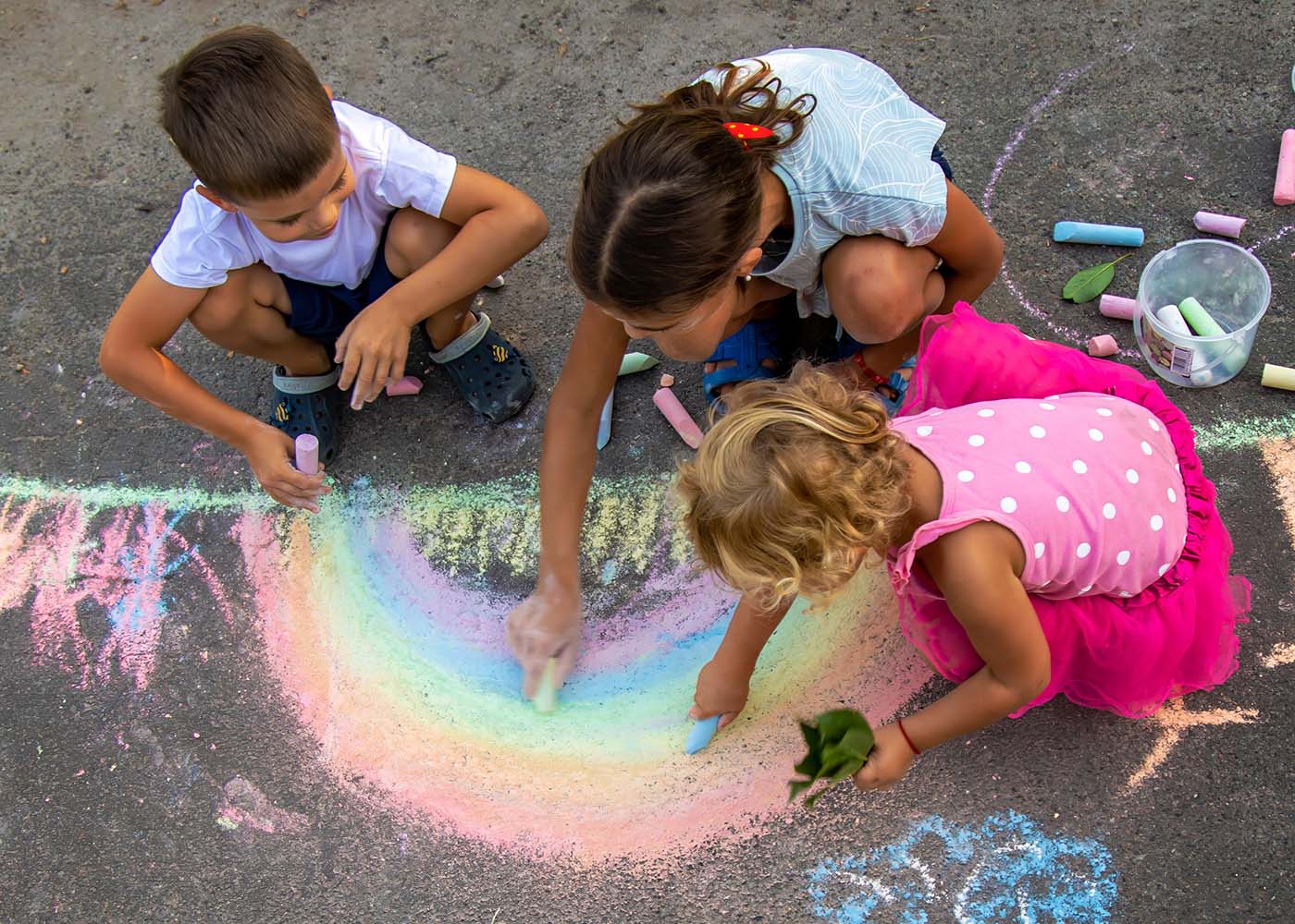 Three children using colorful chalk to draw a rainbow on the concrete sidewalk