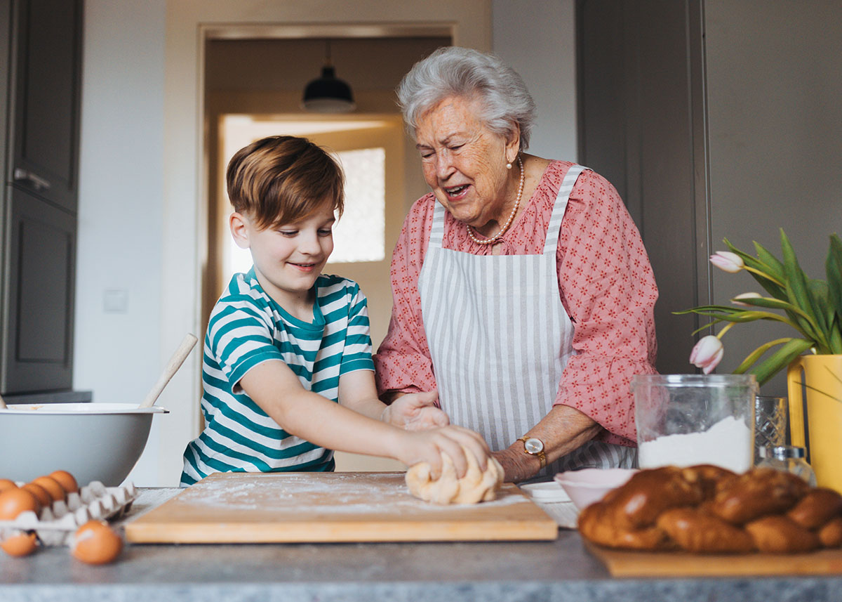 12 year old boy in a turquoise striped shirt kneading dough on a wood dough board 
with his grandmother who is wearing a white apron and salmon house dress