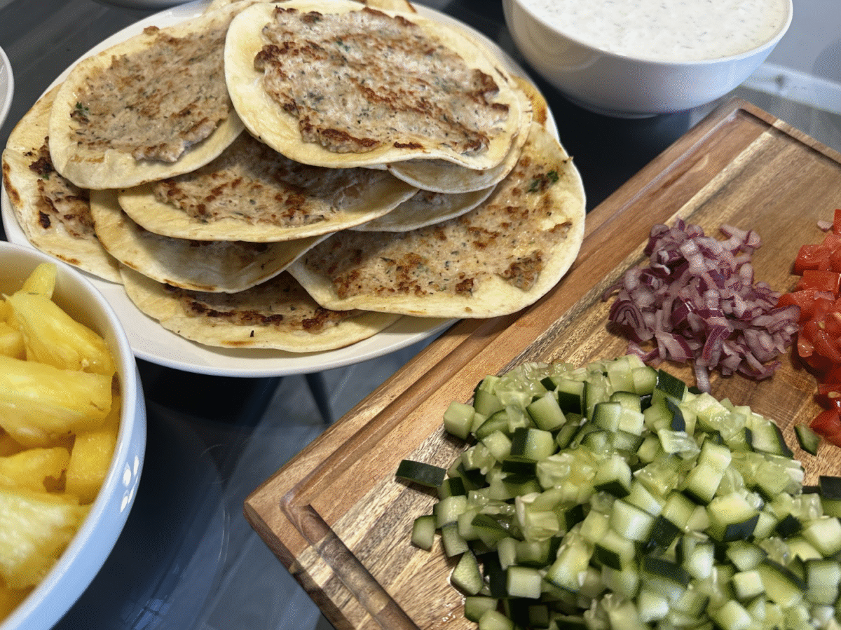 A cutting board topped with diced tomatoes, red onion and cucumbers next to a stack of chicken gyro tacos.