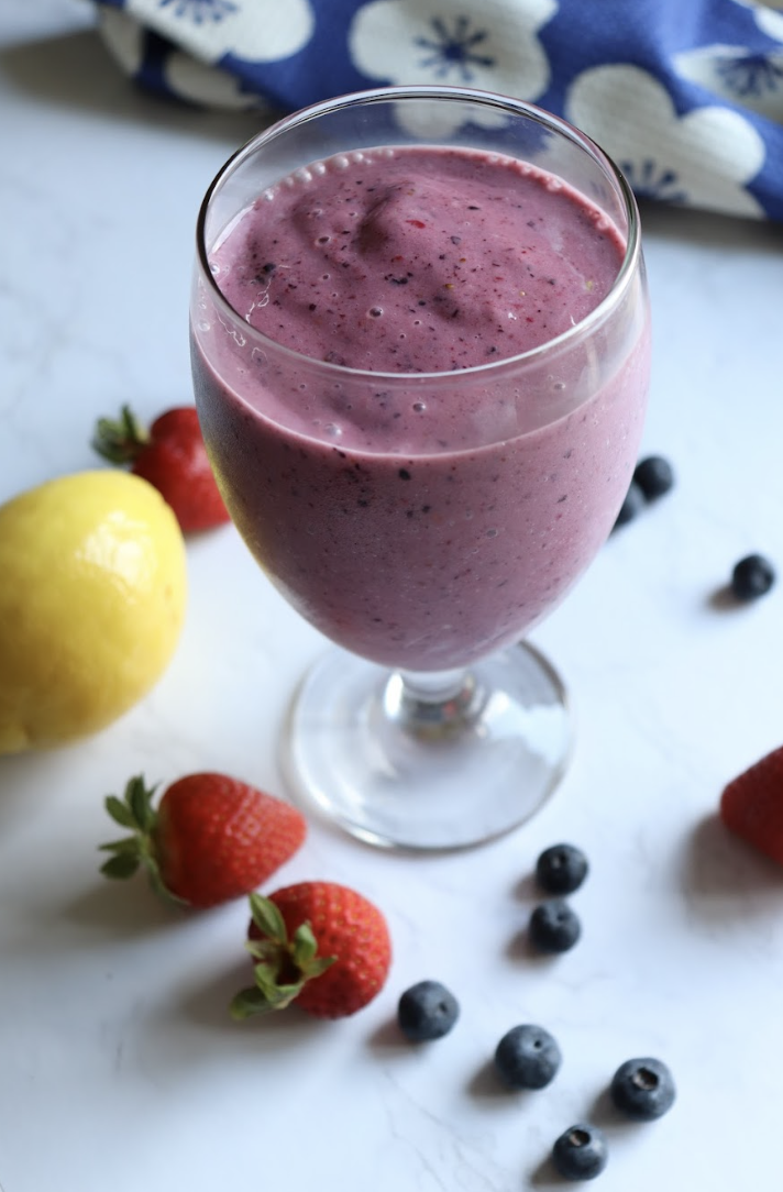 A glass of lemon berry smoothie on a white countertop is spread with fresh berries.
