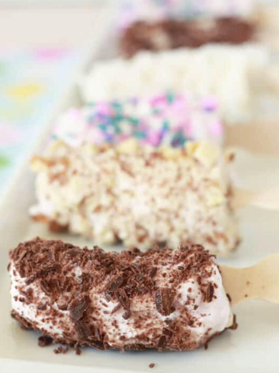 Frozen bananas on a popsicle stick dipped in yogurt and topped with a sprinkles, nuts, coconut, and chocolate flakes.