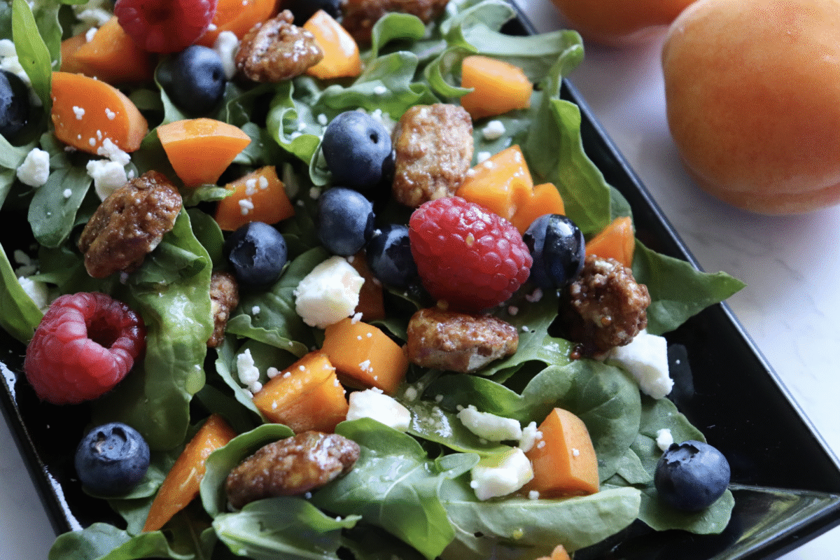 arugula salad with berries and feta on a black serving plate with apricots on the side.