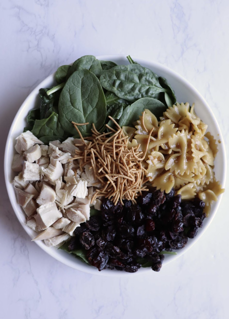 A serving bowl with bowtie pasta, chicken, spinach, chow mein noodles and dried cranberries.