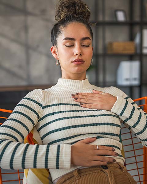 Woman in cream-colored turtleneck sweater with horizontal blue stripes, sitting in a chair, focused on deep breathing as she manages the anxiety of back-to-school