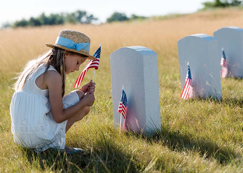 Young girl dealing with the grief of loss by honoring them at their grave, wearing a white sun dress a sun hat and holding an American flag