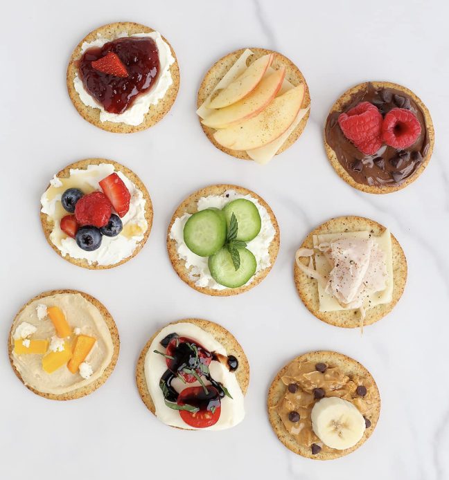 a selection of crackers with different toppings including apple slices, jam, turkey, peanut butter, and cucumber slices.