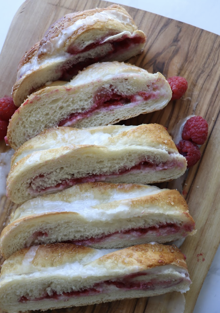 raspberry braided bread topped with a powdered sugar glaze and fresh raspberries on a wooden cutting board and cut into slices. 