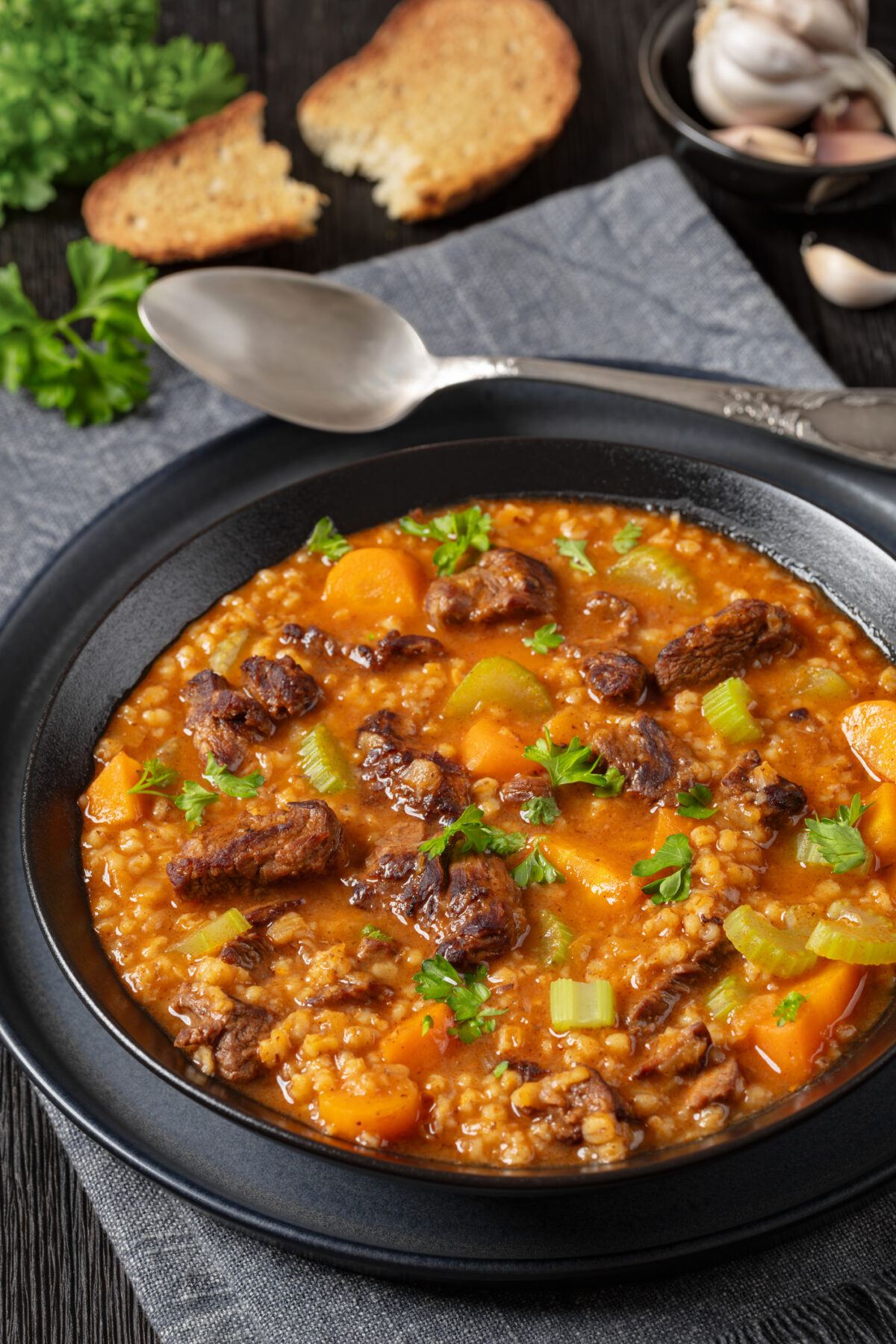 classic beef barley soup with tender chunks of roast beef, fresh veggies and seasoned broth in black bowl on dark oak wooden table with bread and spoon, vertical view, close-up