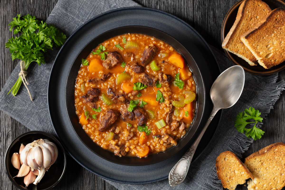 beef barley soup with tender chunks of roast beef, fresh veggies and seasoned broth in black bowl on dark oak wooden table with bread and spoon, horizontal view from above, flat lay, close-up