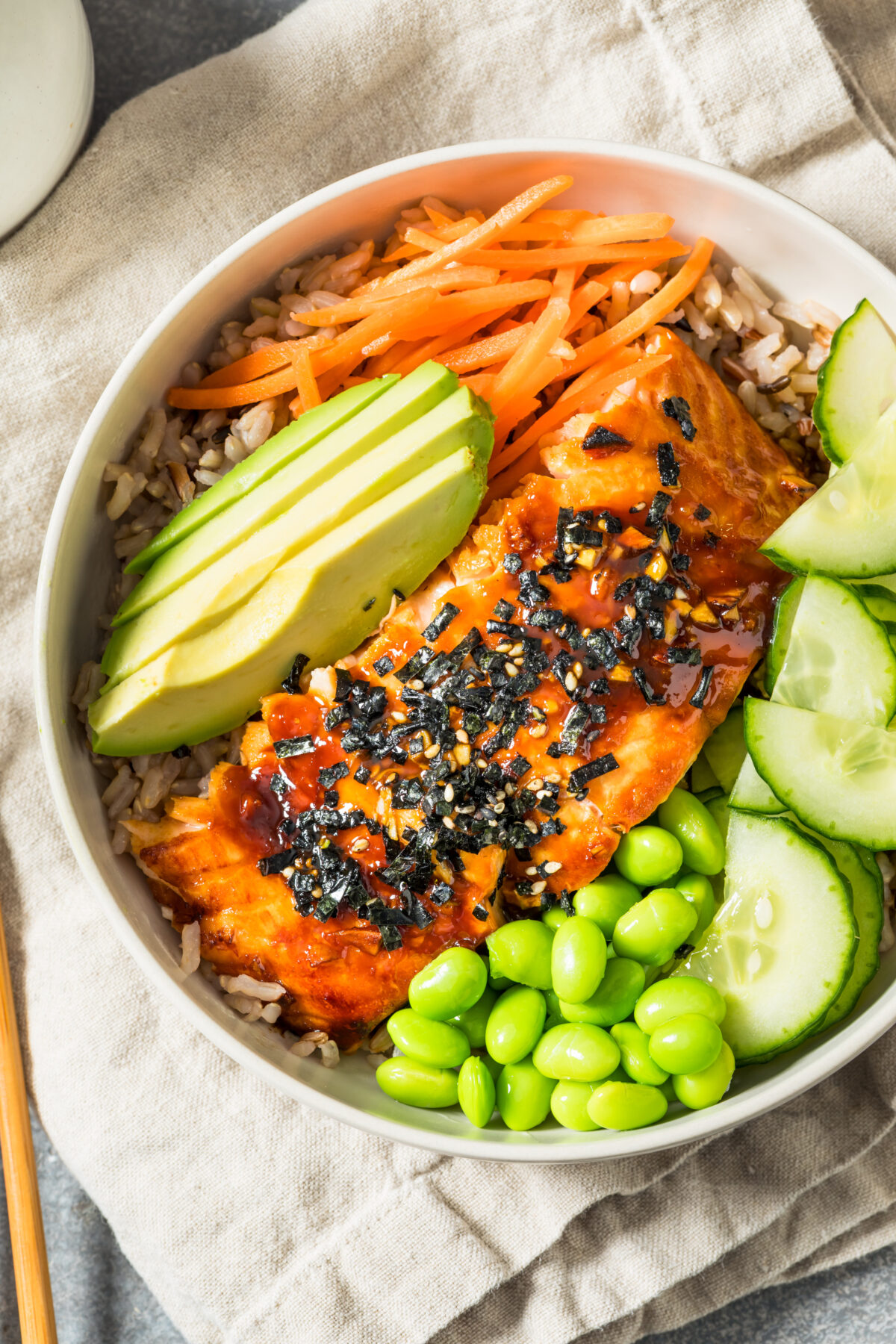 teriyaki salmon bowls topped with cucumber, avocado, carrots and edamame with a linen and chopsticks