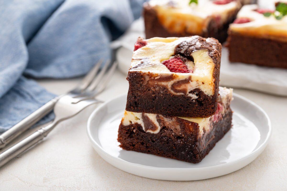cheesecake brownies topped with raspberries on a small serving plate with forks and a blue linen