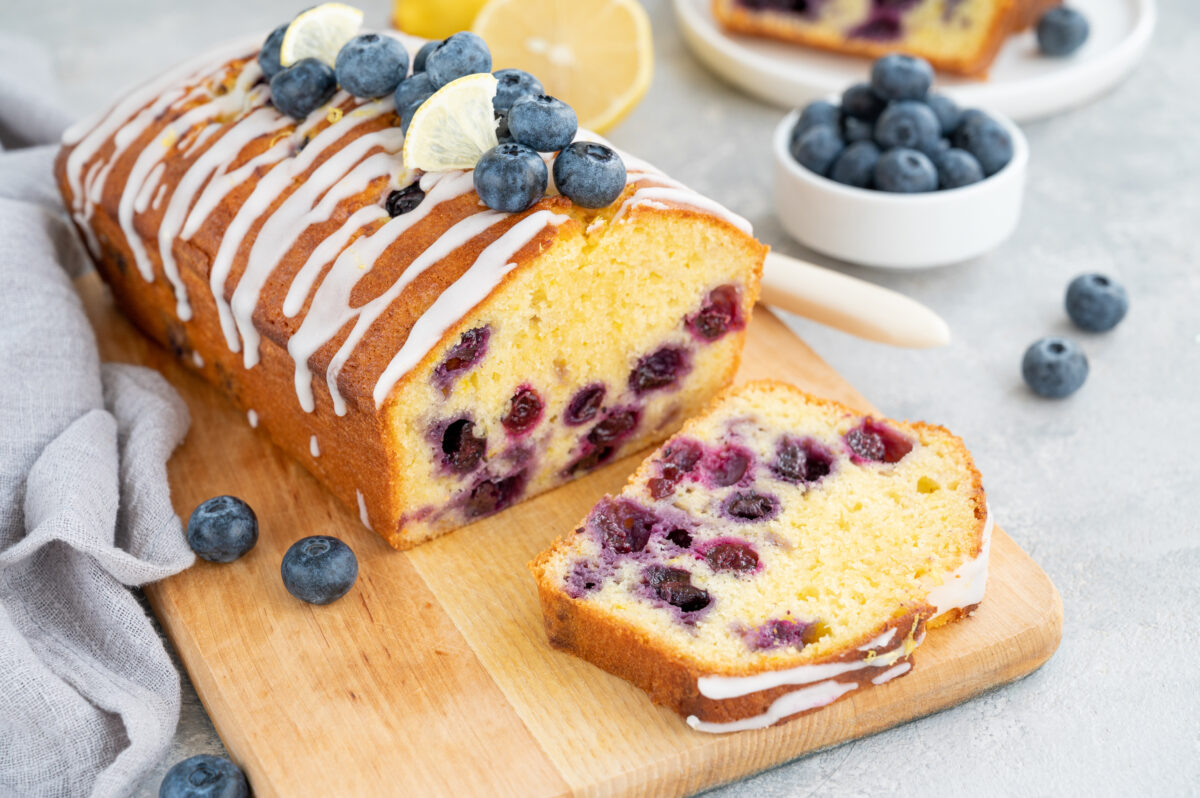 Lemon blueberry cake with lemon icing and fresh berries on top on the board on a gray concrete background with cup of tea.