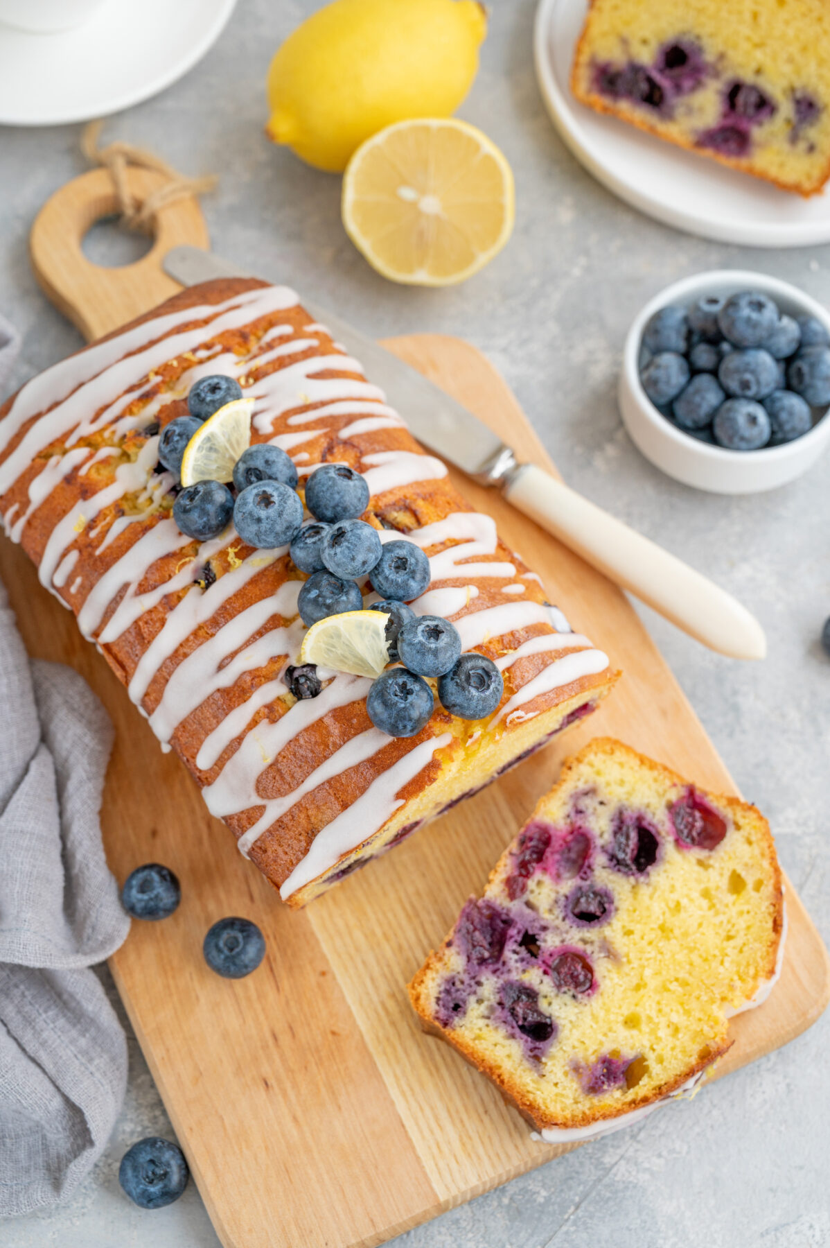 Lemon blueberry cake with lemon icing and fresh berries on top on the board on a gray concrete background with cup of tea.