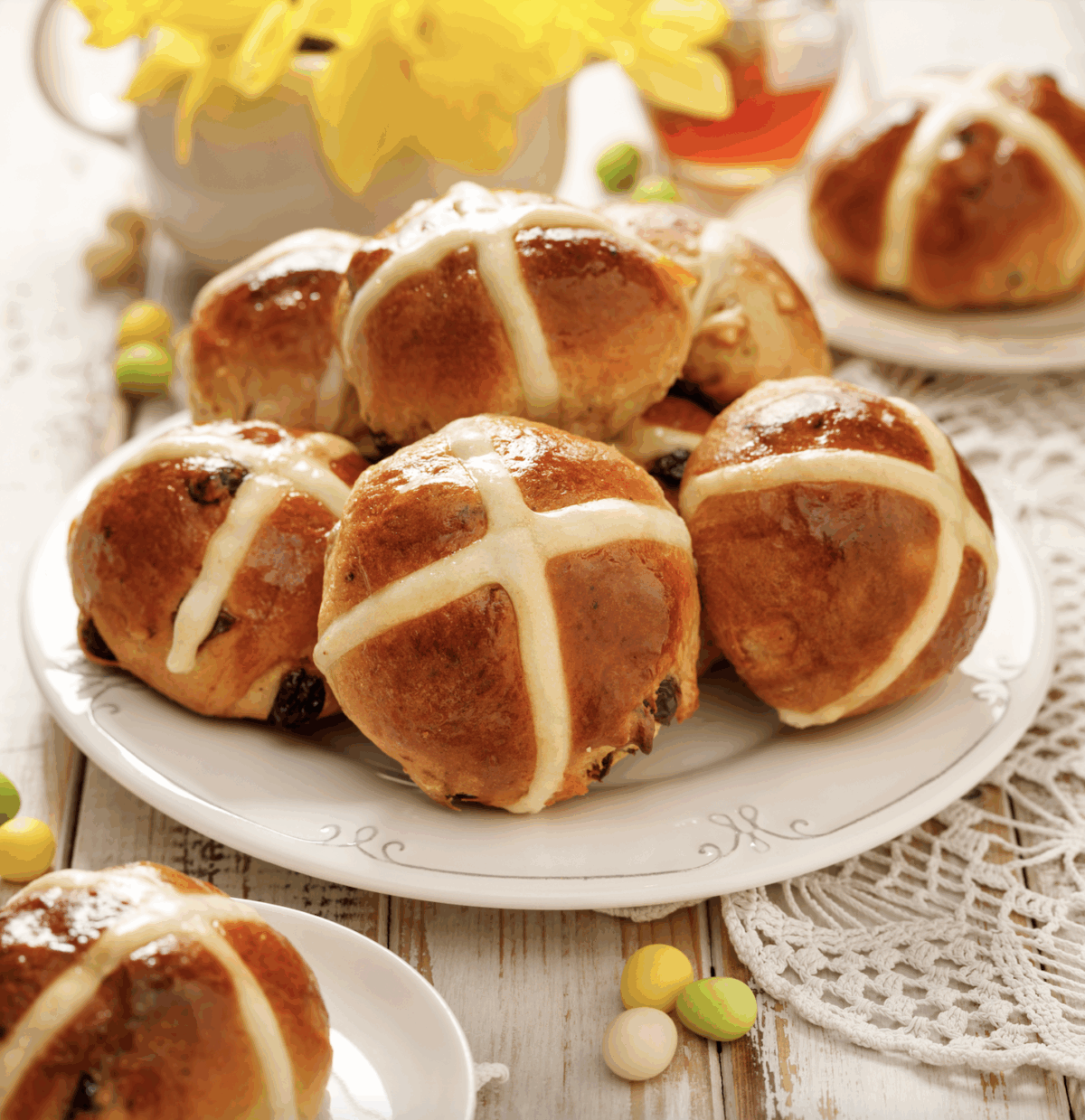 hot cross buns on a serving plate with scattered candy eggs and flowers in the background