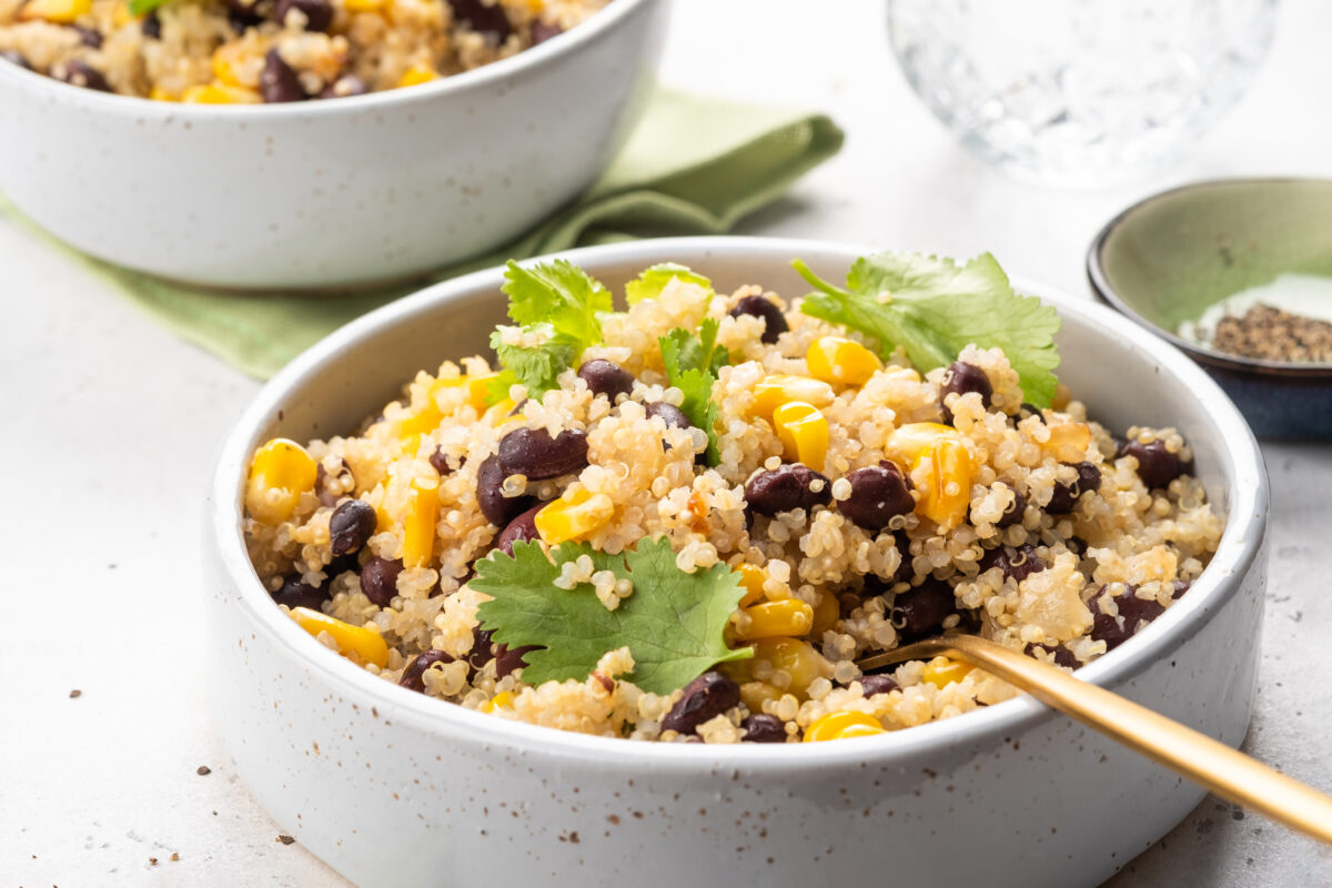 Two bowls contain a quinoa salad mixed with black beans and corn and garnished with cilantro. This bowl sits on a light table during a casual lunch.