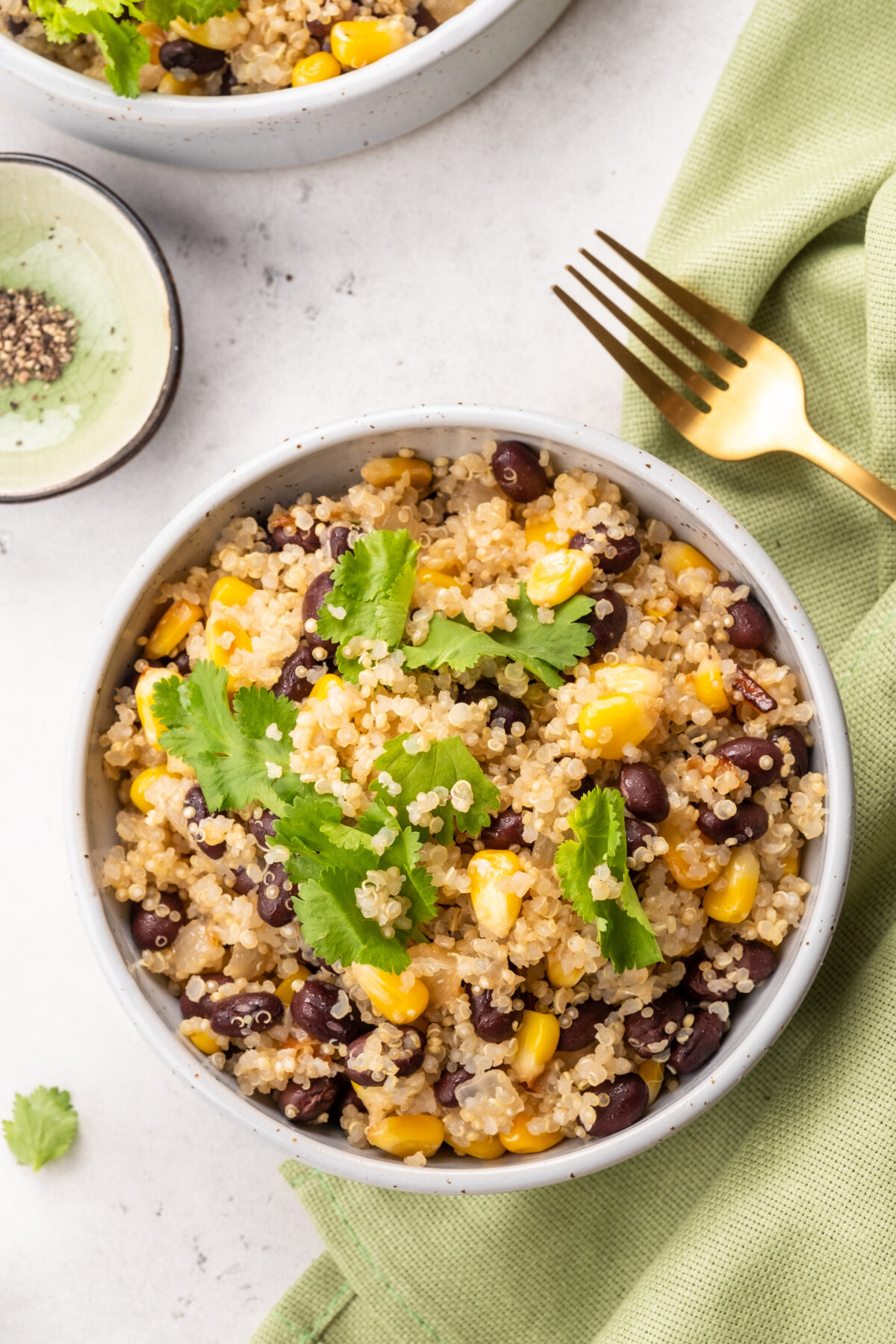 Two bowls contain quinoa salad mixed with black beans and corn, garnished with cilantro. These bowls are placed on a light table during a casual lunch setting.