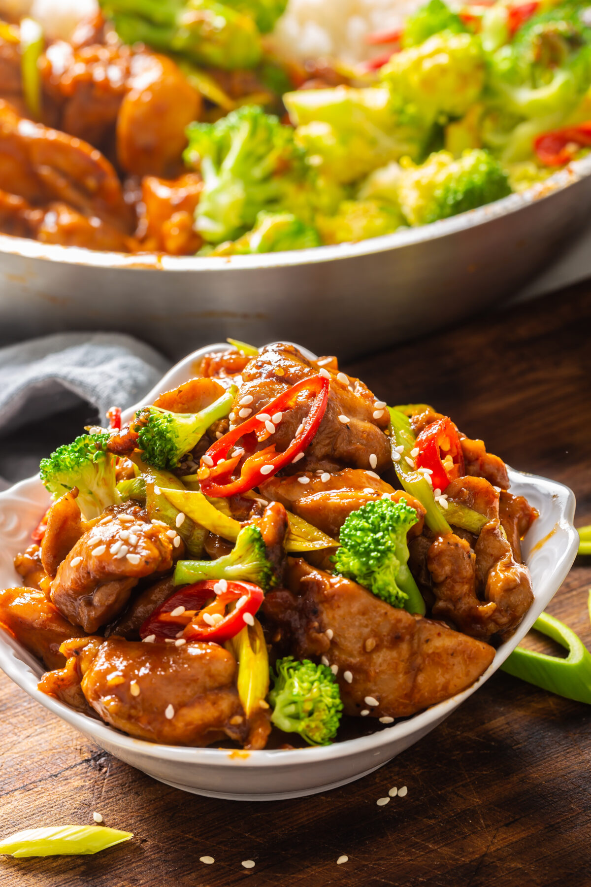 Portrait of a teriyaki chicken bowl sprinkled with seesame seeds, fresh chillies and broccoli with more food in a bowl in the back.