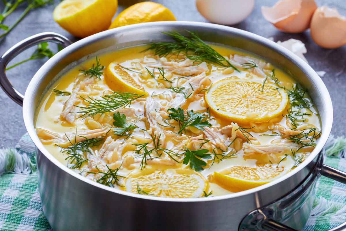 avgolemono - delicious creamy greek chicken soup with lemon and herbs in a casserole on a concrete table with ingredients at the background, view from above, close-up