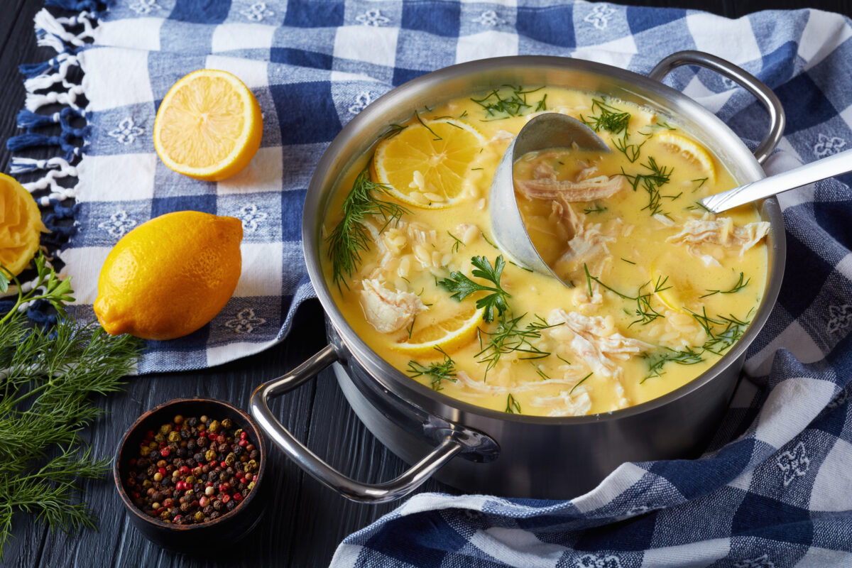 avgolemono - delicious creamy greek chicken soup with lemon, egg yolk, pasta risini and herbs in a casserole with soup ladle, on a black wooden table with kitchen towel, classic recipe, close-up