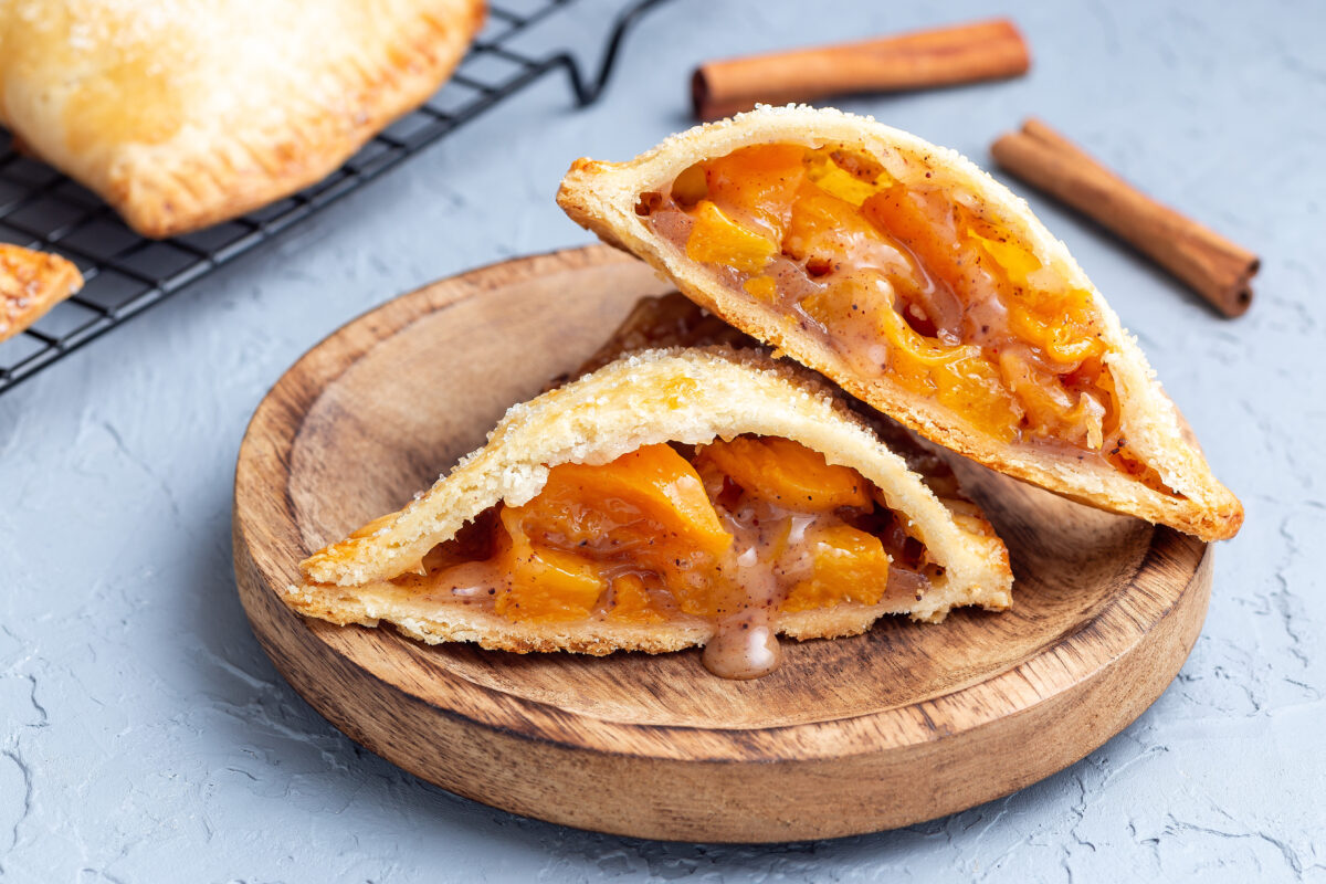 Homemade small peach and cinnamon hand pies with crust dough, sprinkled with sugar, on a wooden plate.