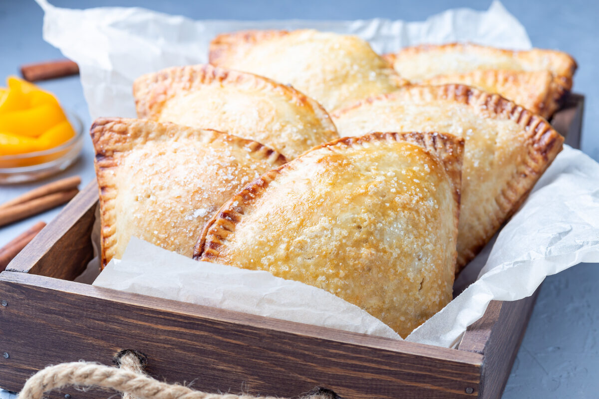 Homemade small peach and cinnamon hand pies with crust dough, sprinkled with sugar, on a wooden tray.