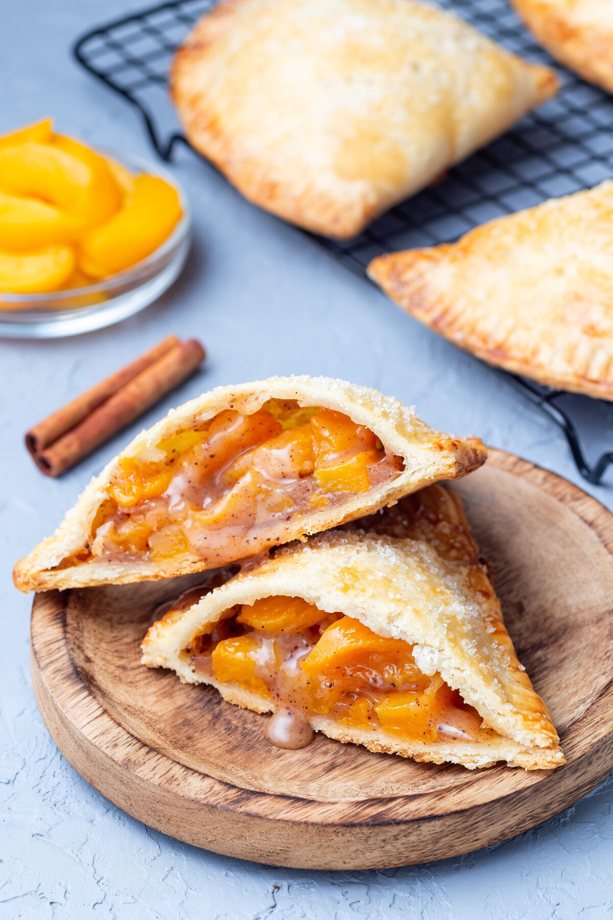 Homemade small peach and cinnamon hand pies with crust dough, sprinkled with sugar, on a wooden plate and cooling rack.