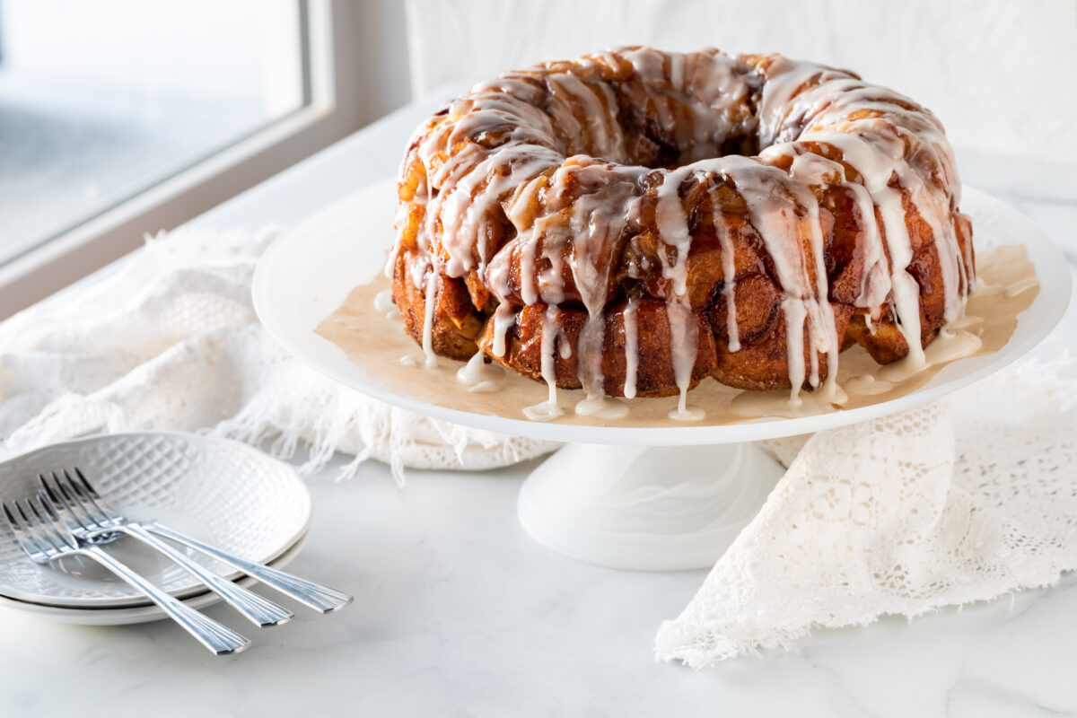 An apple cinnamon pull apart coffee cake drizzled with icing, on a pedestal stand next to a bright window, ready for serving.