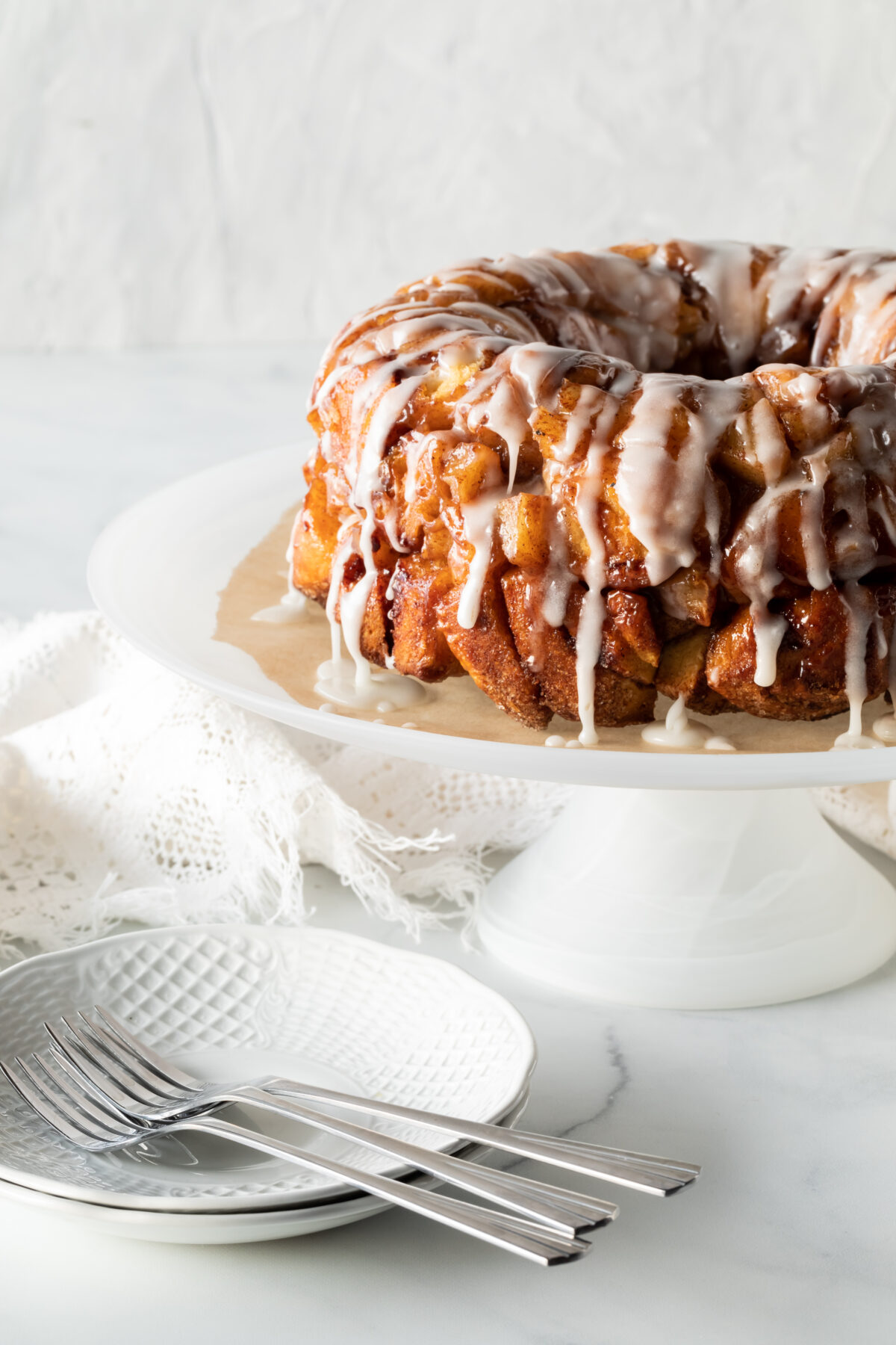 An apple cinnamon pull apart bread cake with icing on a pedestal stand with dishes for serving.