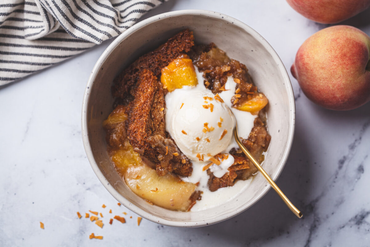Portion of peach cobbler with a scoop of ice cream in a bowl, white marble background, top view.