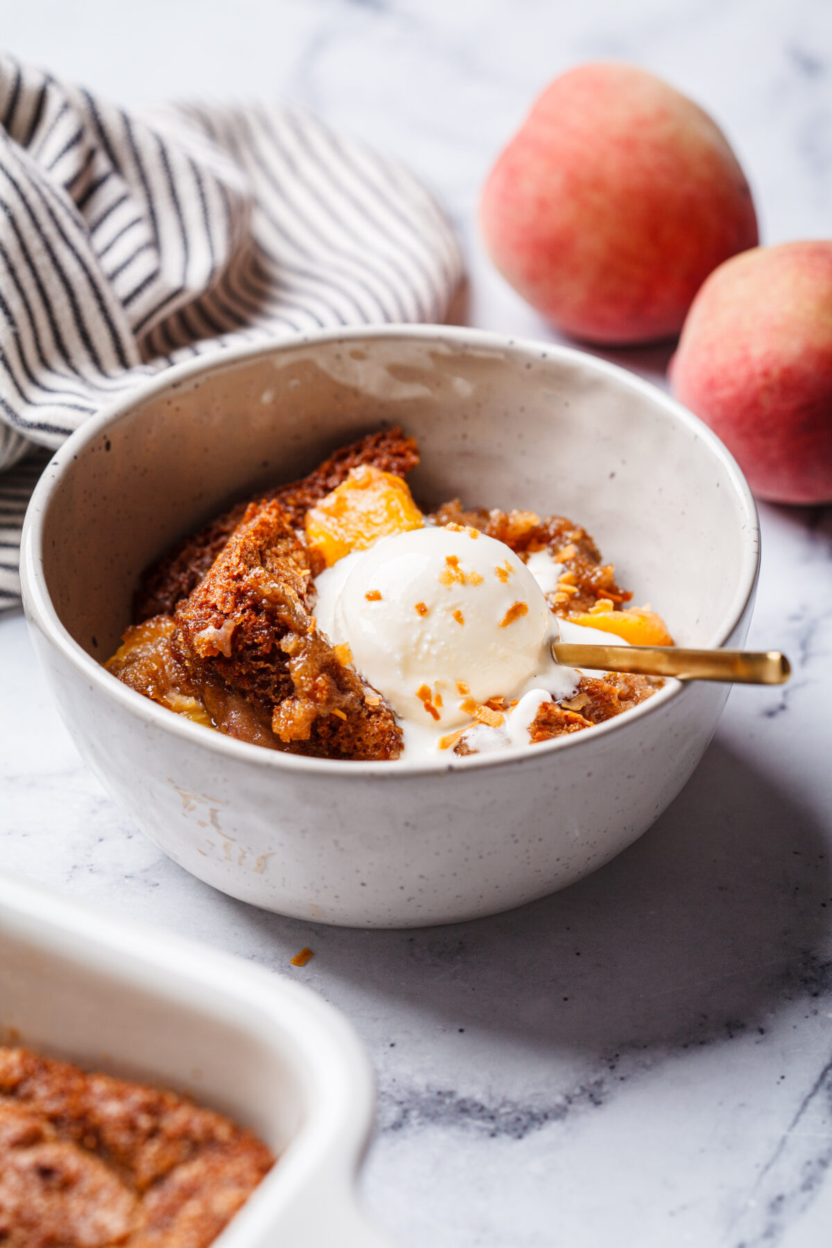 Portion of peach cobbler with a scoop of ice cream in a bowl, white marble background.