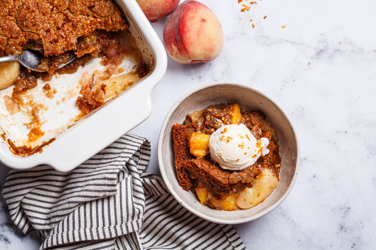 Portion of peach cobbler with a scoop of ice cream in a bowl, white marble background, top view.