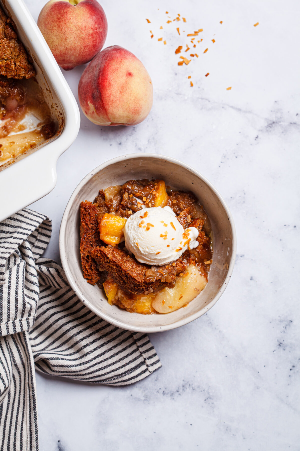 Portion of peach cobbler with a scoop of ice cream in a bowl, white marble background, top view.