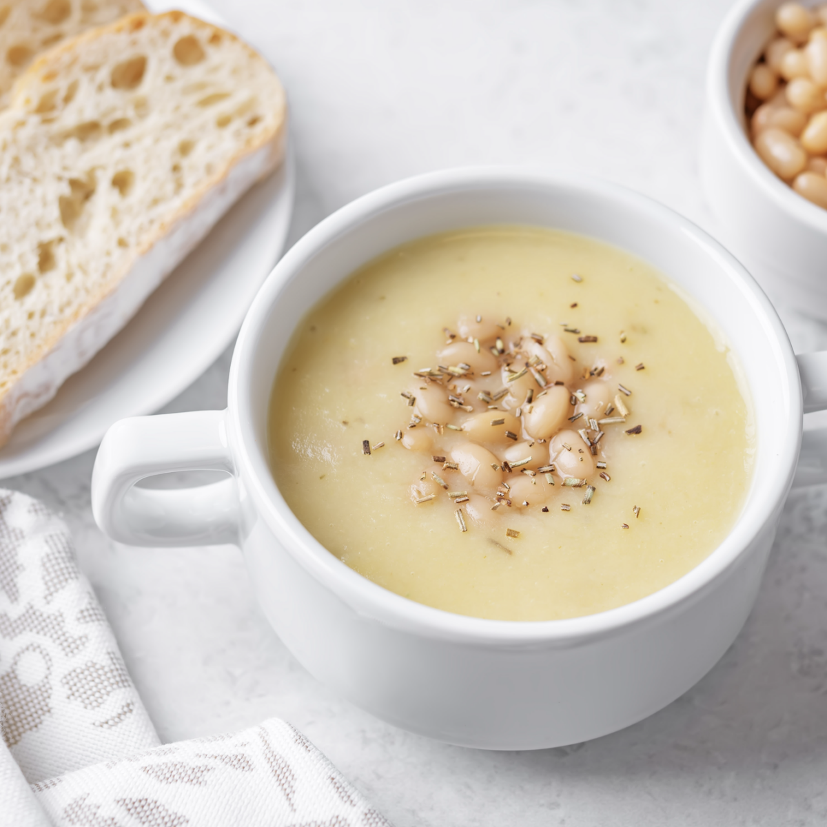 a close up image of white bean soup in a serving bowl topped with fresh rosemary
